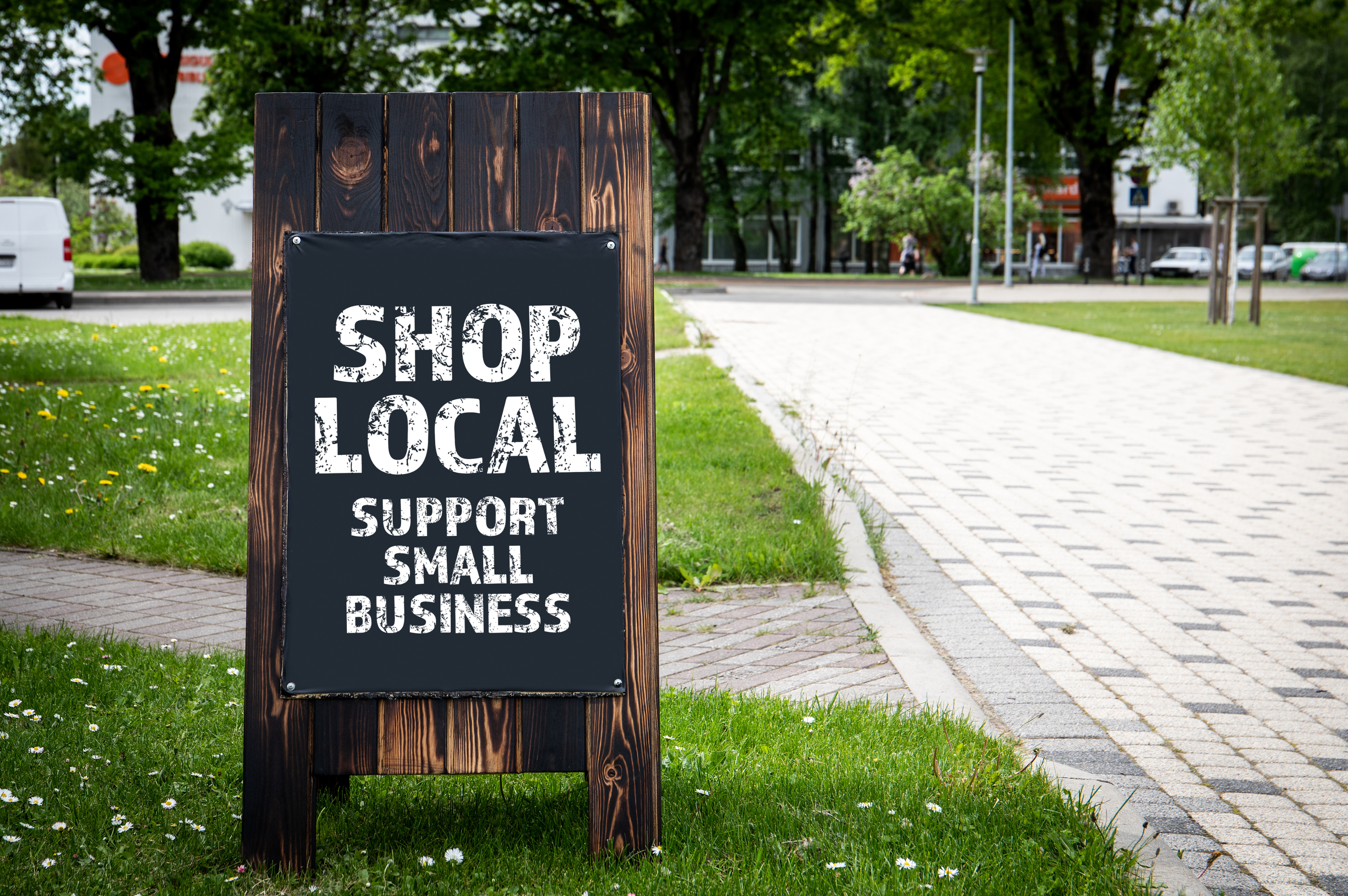 A sidewalk sign encouraging people to shop at local businesses, displayed on a paved walkway with a grassy area and trees in the background